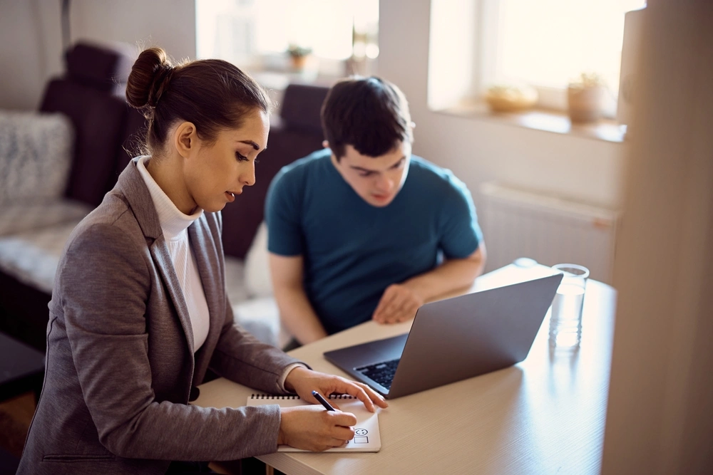 disabled, down syndrome boy working with carer on laptop